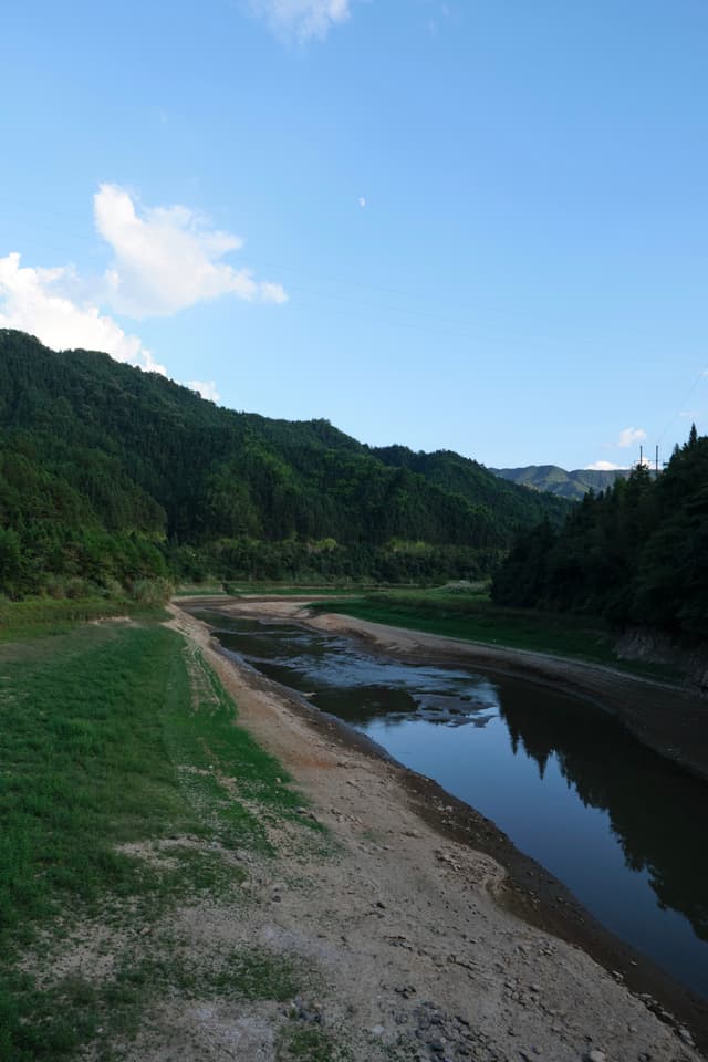 A narrow river with muddy banks flows through a valley flanked by dense green hills under a blue sky with scattered clouds