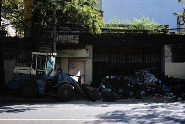 A blue and white front-end loader is positioned next to a large pile of dark, lumpy material on an asphalt road, with a dark wall and green foliage in the background under sunlight