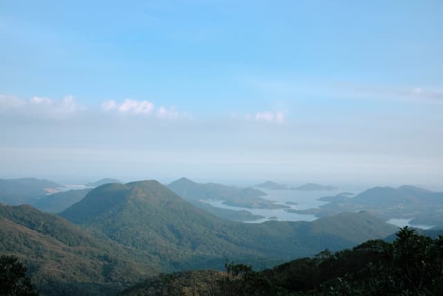 Panoramic view of layered green mountain ranges, hazy distant lake with islands, clear blue sky