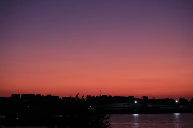 Sky transitions from dark purple to orange-red, silhouetted treeline and distant lights along the horizon, water in the foreground reflects light