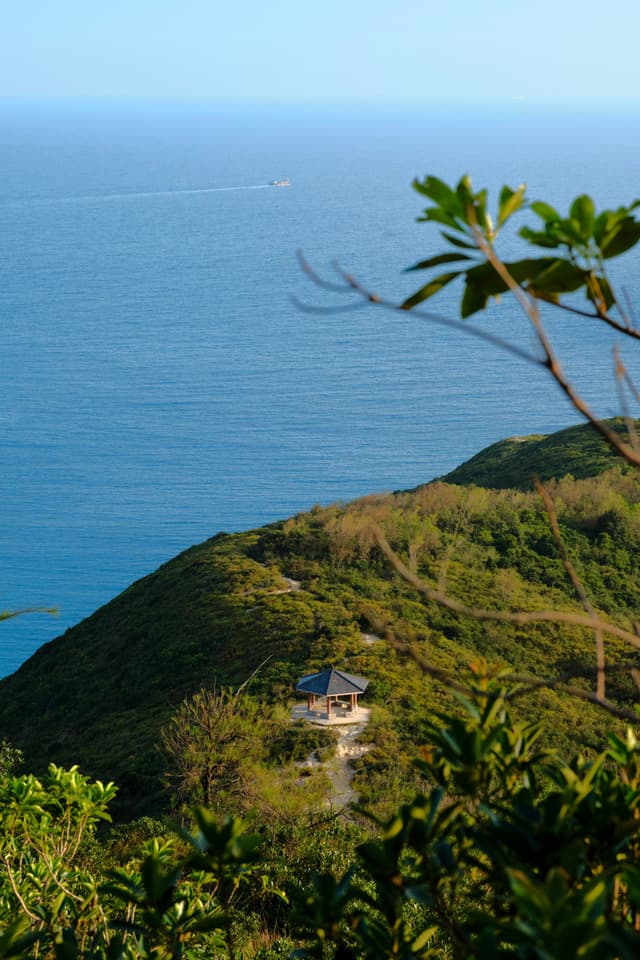 Small structure on green hill overlooking blue ocean, clear sky, foreground leaves