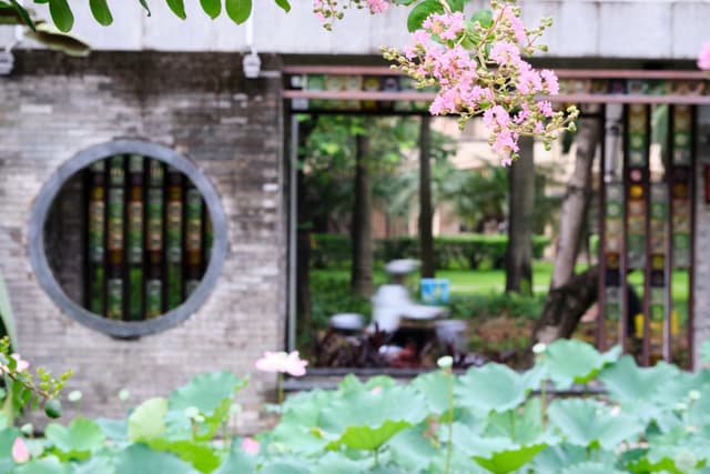 Traditional garden wall with circular window and open doorway revealing blurred figure in green courtyard, lily pads in foreground, pink flowers overhead