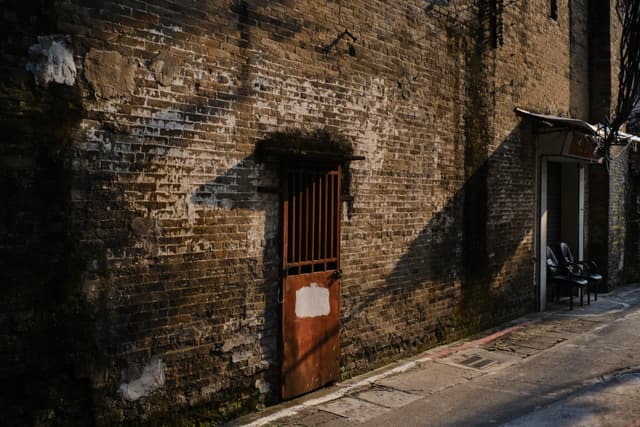 Weathered brick wall in a dim alley, featuring a rusty red metal gate and a diagonal shadow. A person sits in a recessed doorway on the right