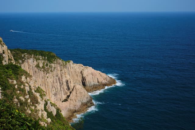 Rugged cliff face beside dark blue ocean under clear sky, small boat in distant upper left