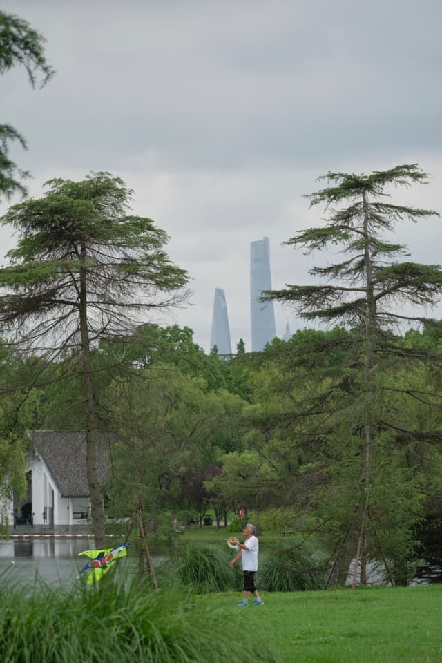 Person playing badminton in a park; tall pine trees frame a white building and distant modern skyscrapers under a cloudy sky