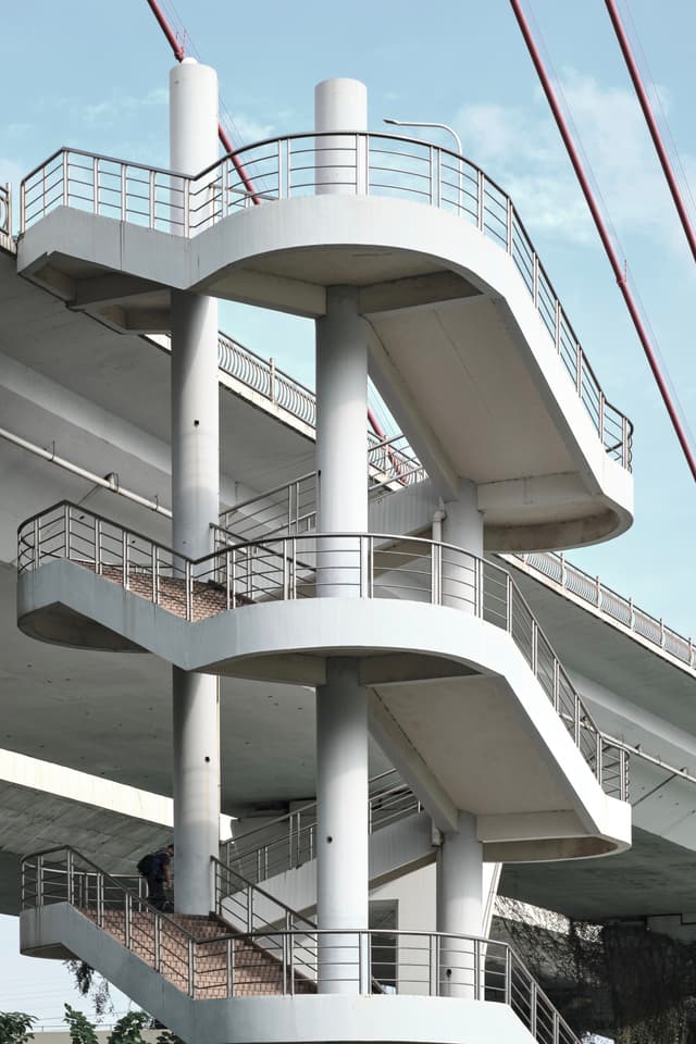Multi-level concrete spiral pedestrian walkway beneath a bridge, featuring white pillars, metal railings, and grey stairs. A person is visible on the lowest steps. Red suspension cables are in the background against a light blue sky
