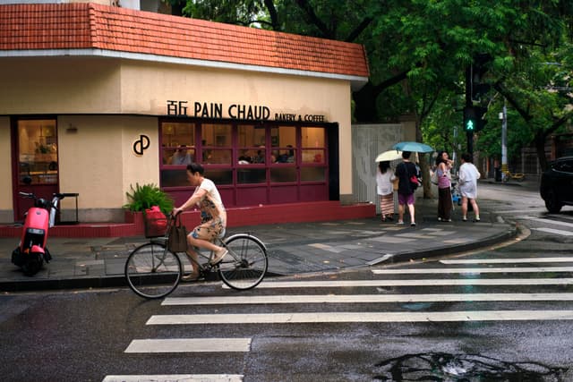 Cyclist with basket of greenery crosses a wet street on a crosswalk in front of a Pain Chaud bakery, with pedestrians holding umbrellas on the sidewalk