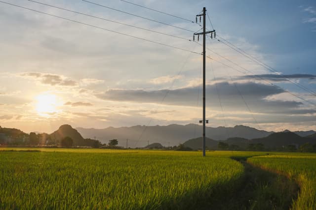 Green field with tall utility pole, setting sun, mountains, and cloudy sky