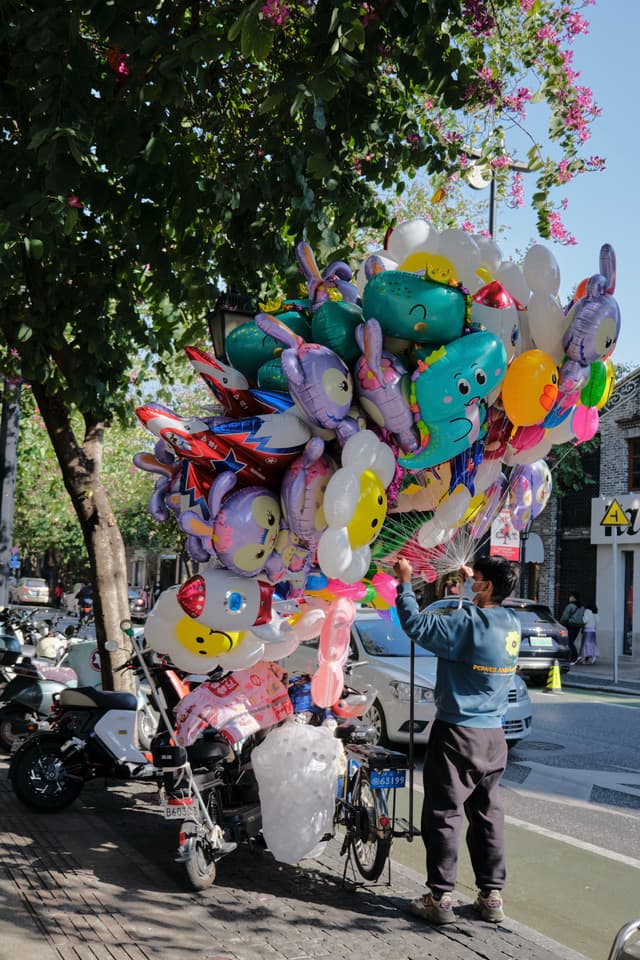 A person stands next to a large cluster of multicolored, character-shaped balloons attached to a cart on a sunny outdoor street. Parked scooters and trees are visible in the background