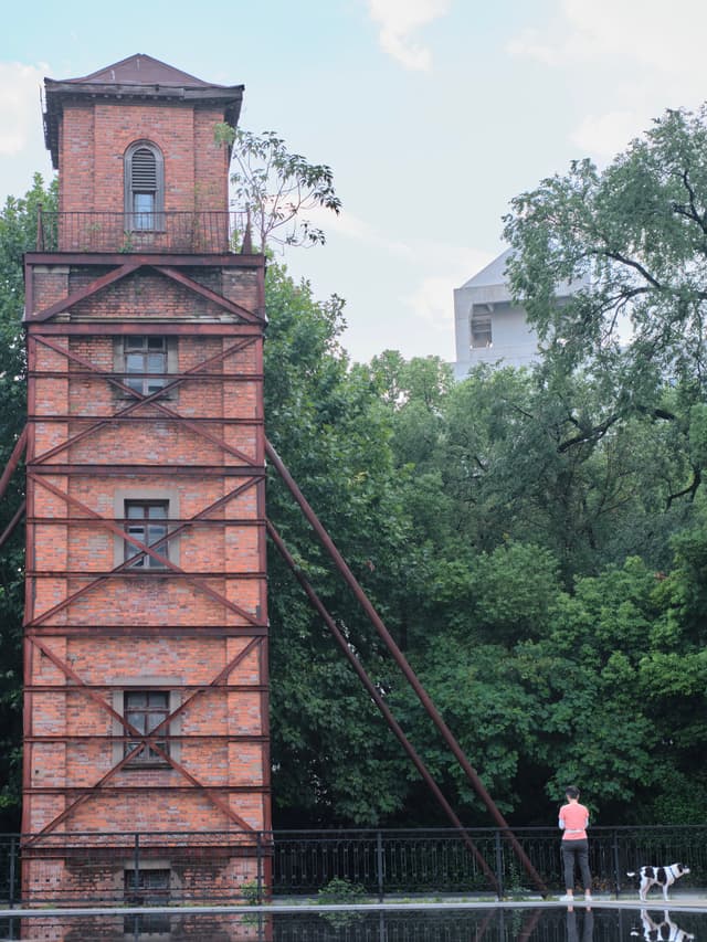 Tall red-brick tower with external metal frame, green trees, person in orange shirt on reflective surface, distant white building