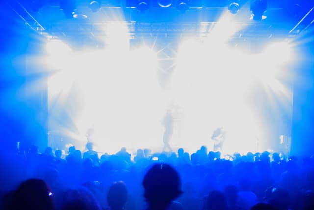 Silhouetted audience faces brightly lit stage with performers amidst blue and white spotlights