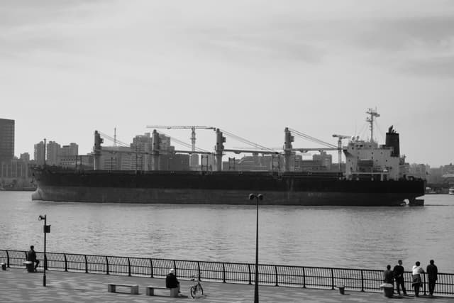 Black and white photograph of a large cargo ship on a river, with a city skyline in the distance and a waterfront promenade with people in the foreground